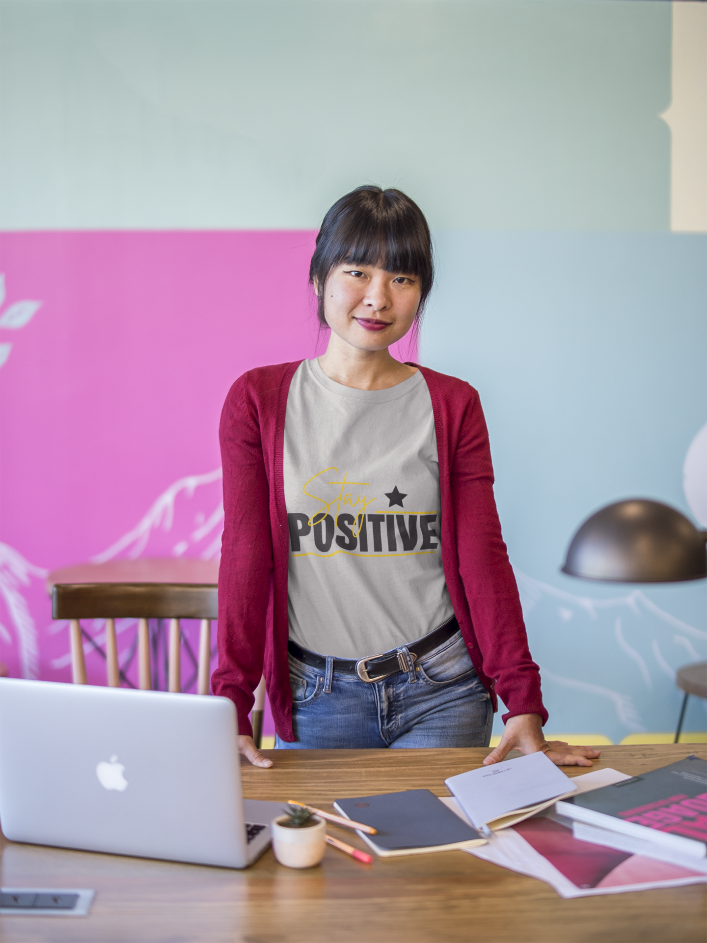 Person standing behind a desk with a laptop and books, against a colorful wall.