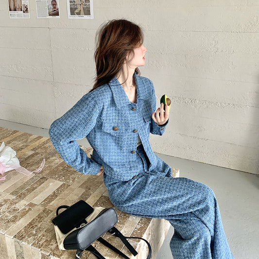 A woman sitting on a bench, wearing a blue coat with long sleeves and matching wide-leg pants, holding a mobile phone in her right hand.